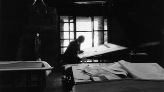 American architect Frank Lloyd Wright  working on designs while sitting in front of a drafting table in his studio at...