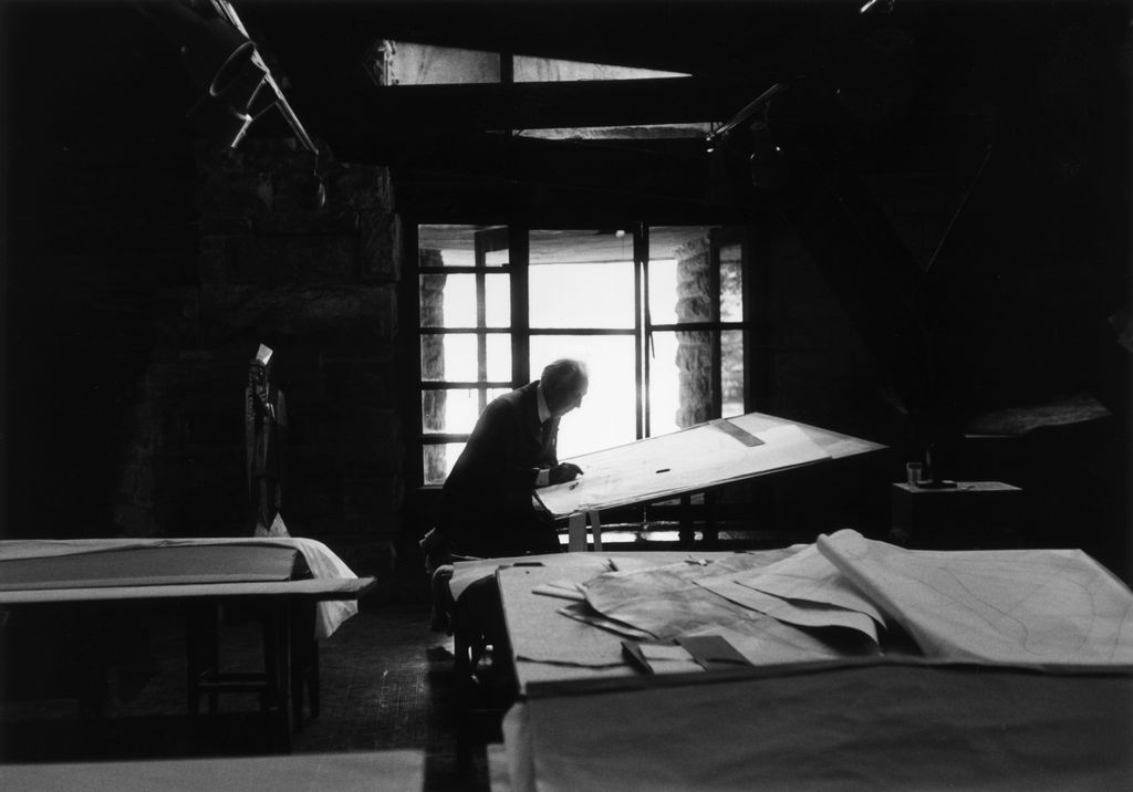 American architect Frank Lloyd Wright  working on designs while sitting in front of a drafting table in his studio at...